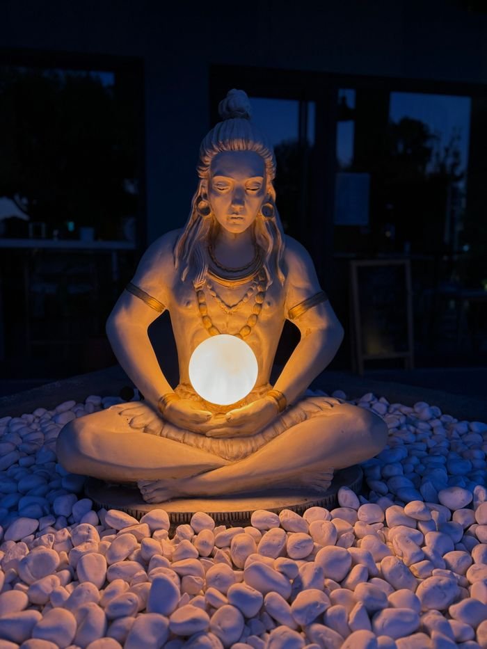 A serene sculpture of Shiva holding an illuminated orb, surrounded by pebbles.